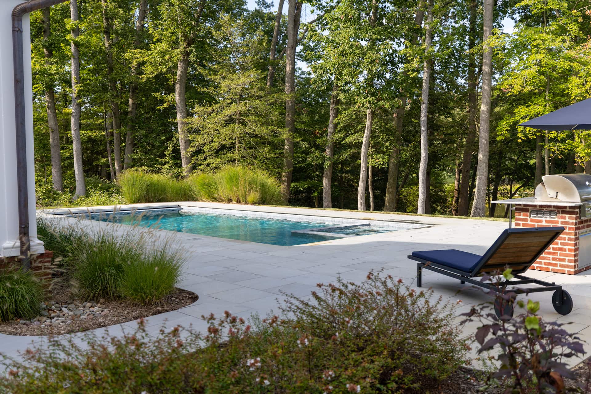 angled view of pool and spa and outdoor kitchen with large trees background