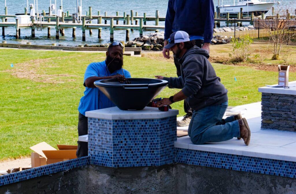 Workers installing a black fire bowl on a blue-tiled poolside structure